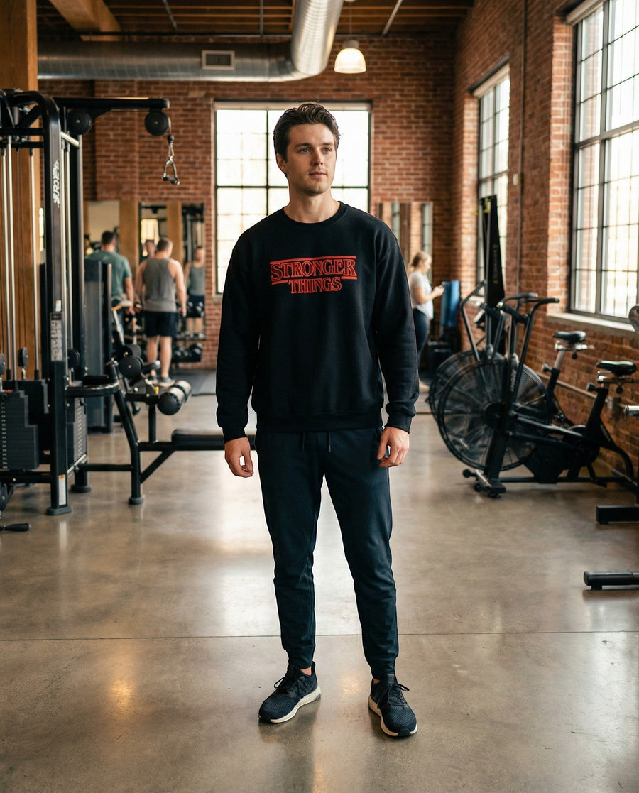 DailyThreadCo. black crewneck sweatshirt with red "Stronger Things" logo embroidery, worn by a man in a gym.