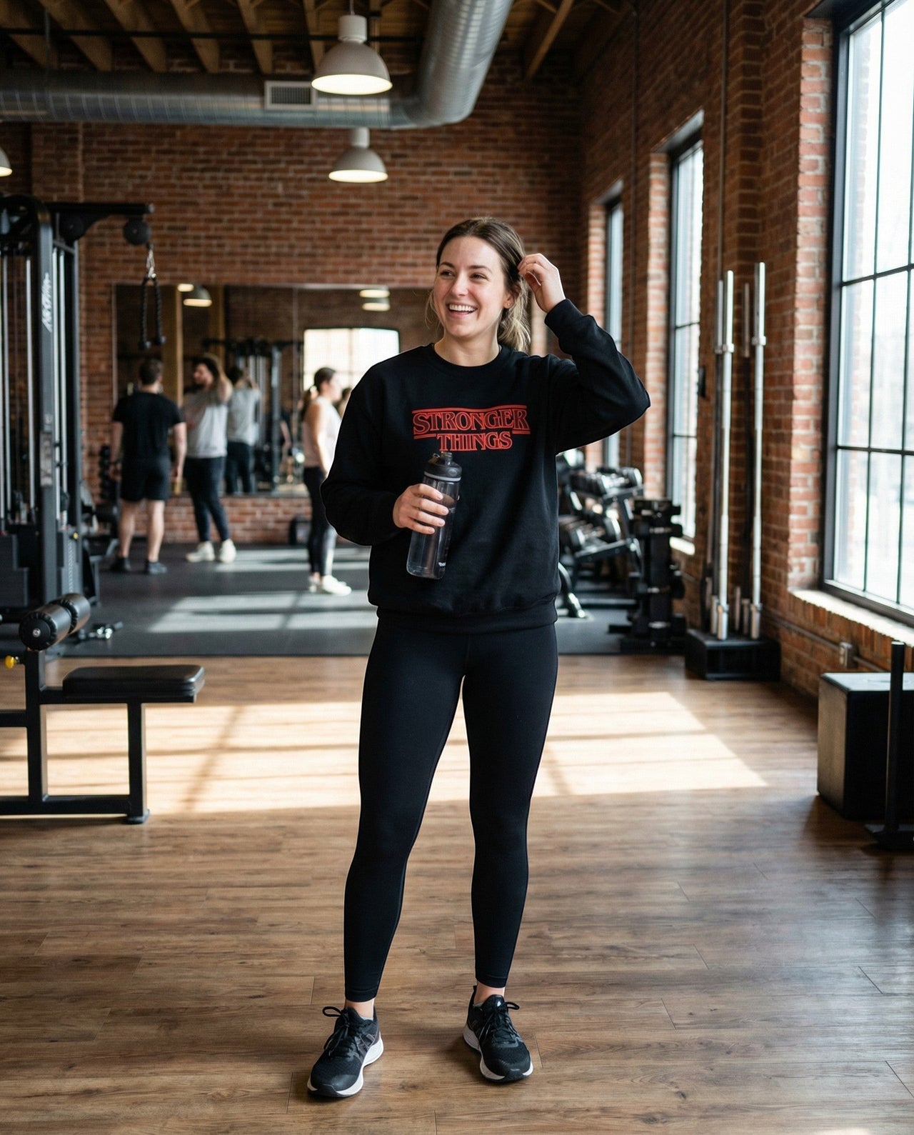 DailyThreadCo. black crewneck sweatshirt with red "Stronger Things" retro logo, worn by a woman in a gym.